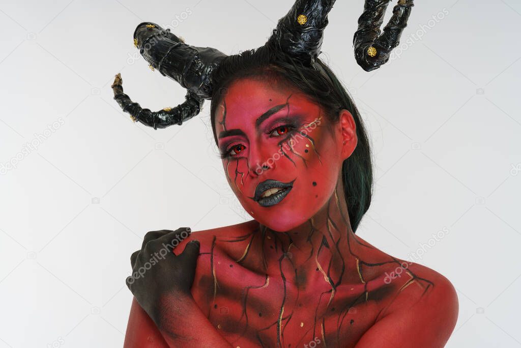 young pretty girl doing body painting characterized by demon with big black horns and red and black body doing poses in photo studio