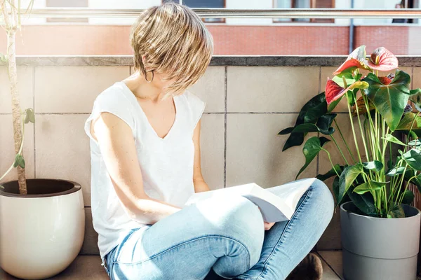 blonde woman reading a book sitting on the balcony floor surrounded by plants at sunset and confined to her home because of the coronavirus pandemic