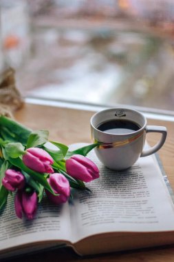 Pencerede koyu, lezzetli, aromatik bir kahve, ilginç bir kitap ve pembe laleler var. Sabah güneşi bir kitabın ve aromatik kahvenin üzerinde parlıyor ve yakınlarda güzel pembe laleler uzanıyor.
