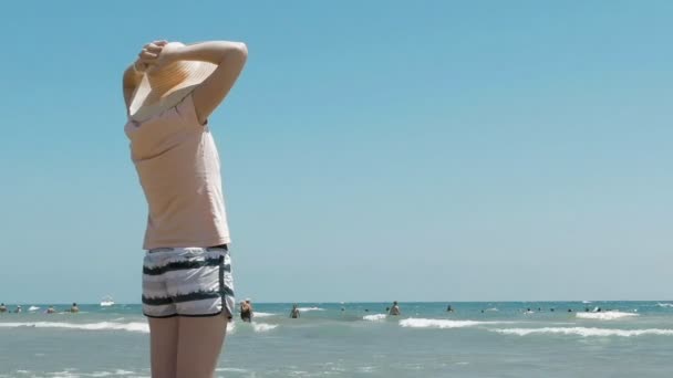Женщина стоит на пляже в джутовой шляпе — стоковое видео