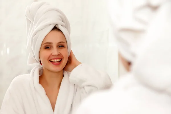 Woman of Slavic appearance in dressing gown smiles in mirror. A happy lady looks in bathroom mirror, touching healthy skin of her face. Gentle and cheerful girl after shower at mirror