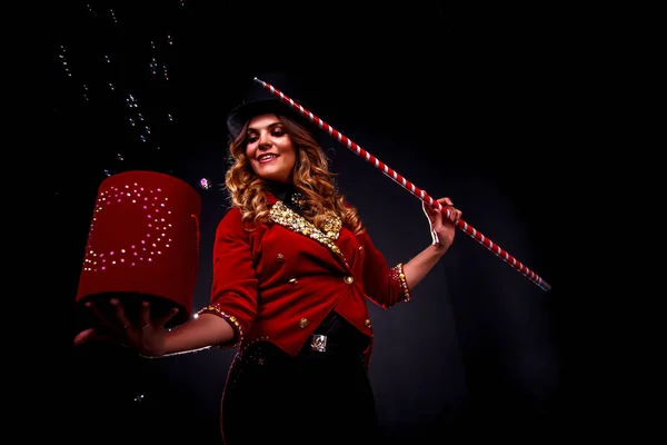 Girl in stage costume and top hat on her head. Female magician, an illusionist in theatrical clothes, with cane, box and soap bubbles in hand on black background. Concept of theatrical performance