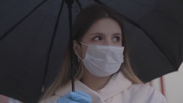 傘の下で医療用マスクやゴム手袋の女の子 悪天候で路上でマスクをした女性 若い女性のスラブの外観のクローズアップ ウイルス対策の概念 Covid 19パンデミック — ストック動画