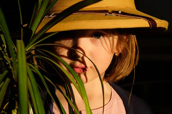 little girl with a short haircut, in a hat, through a magnifying glass looks at the green leaves of a flower in the jungle