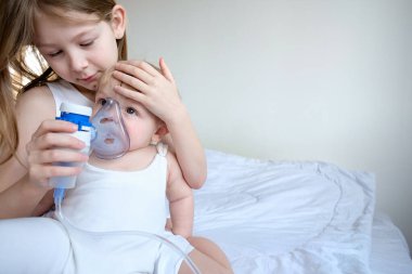 Çocuklar hastalandı. Ablası erkek kardeşine bakıyor ve solunum için bir Gmu maskesi tutuyor. Evde ilaç var. şık beyaz oda