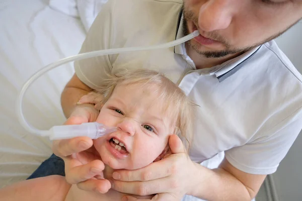 in a white room on his fathers lap sits a child with snot. Dad and son treat the flu, and dad cleans his sons nose with snot