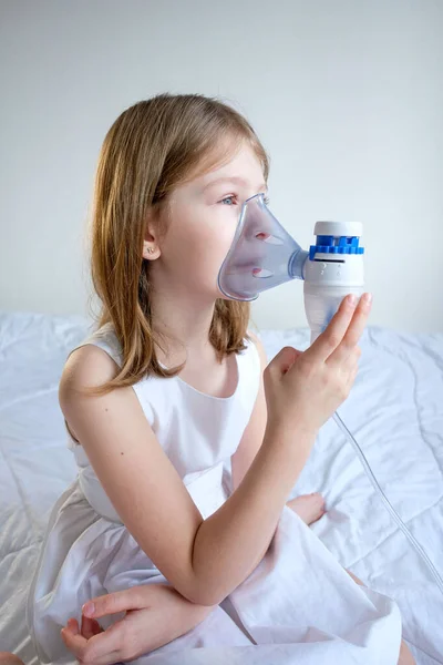 a little sick girl in white clothes sits on a white bed and breathes through an inhaler mask to treat a throat