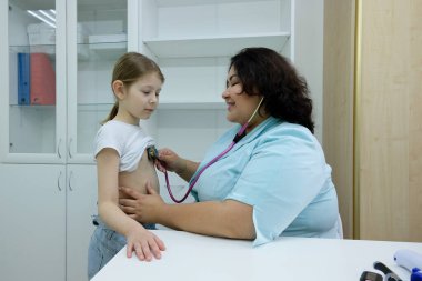 Doktor, ofisteki statoskopla hastanın kalp atışlarını ve nefes alışlarını dinliyor. Küçük kız acil tıbbi muayene