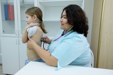 Doktor, ofisteki statoskopla hastanın kalp atışlarını ve nefes alışlarını dinliyor. Küçük kız acil tıbbi muayene