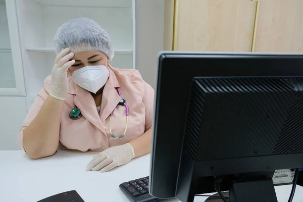 Koruyucu maskeli ve eldivenli bir doktor masasına oturup koronavirüs salgını sırasında resepsiyona giriyor..