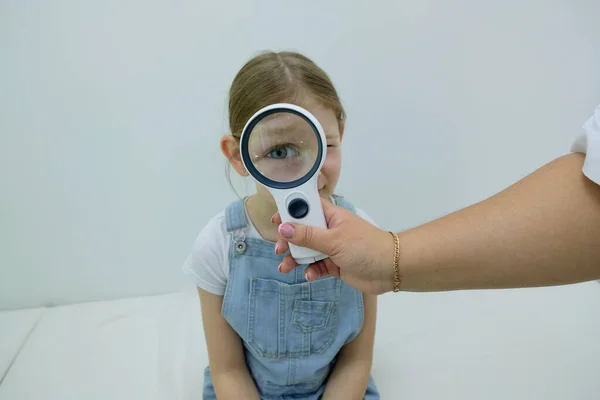 the girl at the doctors office sits on the couch and checks her vision through a magnifier. The baby looks through the magnifying glass and she has a big eye
