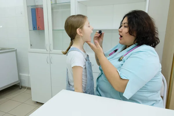 这个女孩带着耳镜去看儿科医师 用舌头抑制剂检查小孩喉咙的时候 笑着耳鼻喉科医生看着小孩 — 图库照片