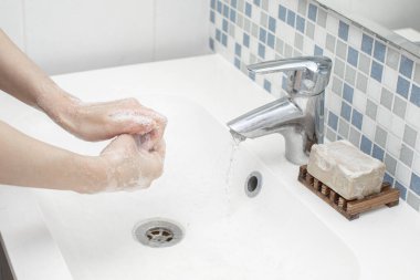 Beyaz kadın elini beyaz lavabosunun önünde köpüklü yeşil sabunla yıkıyor. Onun görüntüsü aynaya yansıyor ve su gümüş musluktan akıyor. Duvarda küçük bir kiremit var..