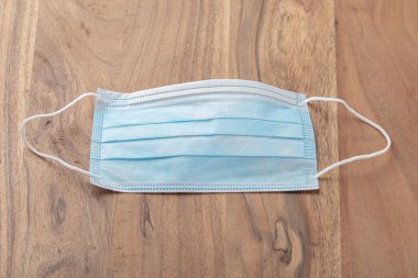 Katı kahverengi meşeden yapılmış ahşap bir masaya yerleştirilmiş mavi bir cerrahi maskenin fotoğrafı. Mavi yüzlü maske yukarı bakıyor ve kullanılmaya hazır..