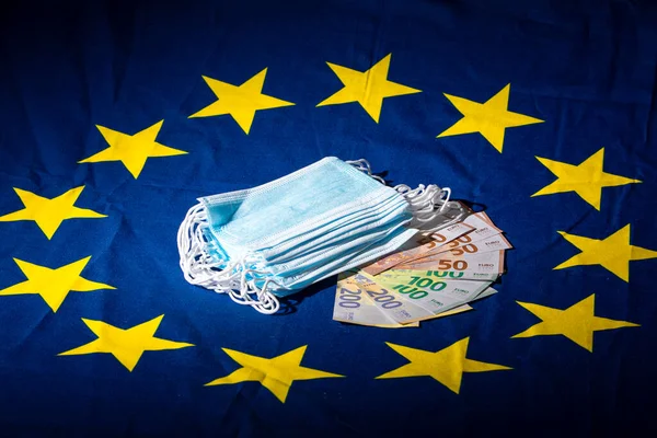 stack of blue surgical masks placed on a stack of banknotes on a European flag