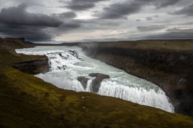 İzlanda 'nın güneybatısındaki en büyük şelalelerden biri olan Gullfoss' un genel manzarası. Gökyüzü bulutlu kraliyet mavisi. Yüzeyden yükselen sisi ve yukarıdaki gökkuşağını ayırt edebiliriz.