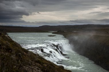 İzlanda 'nın güneybatısındaki en büyük şelalelerden biri olan Gullfoss' un genel manzarası. Gökyüzü bulutlu kraliyet mavisi. Yüzeyden yükselen sisi ve yukarıdaki gökkuşağını ayırt edebiliriz.