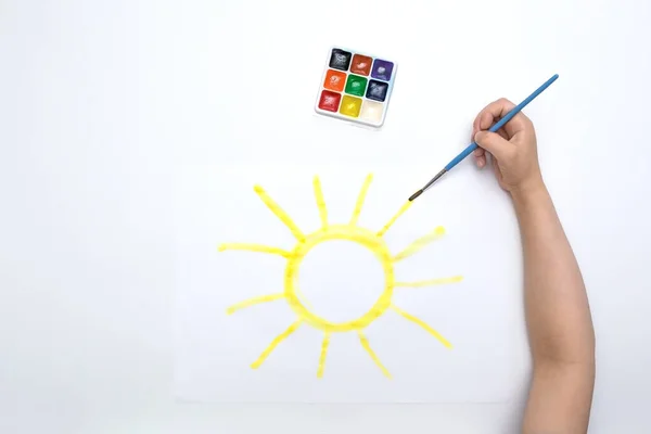 A child\'s hand draws the sun with a brush and paints on a white background
