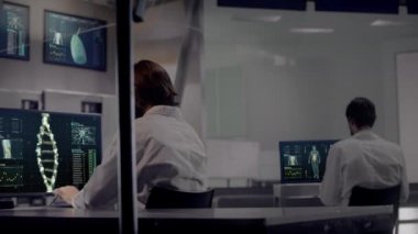 Tıbbi Genetik Mühendisliği Araştırma Biyoteknoloji Laboratuvarı 'nda çalışan iki profesyonel erkek bilim adamı arkadan görüldü. DNA Strand molekülleri ve İnsan Anatomisi grafiklerini gösteren bilgisayarlar.