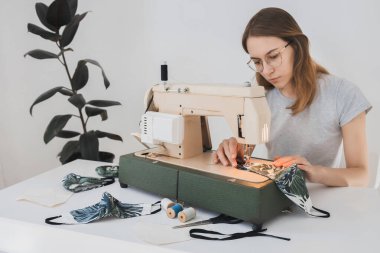 Gribi önlemek için evdeki dikiş makinesine koruyucu ilaç maskesi takan bir kız. Covid-19 beyaz arka plan ev işyeri olan odada.