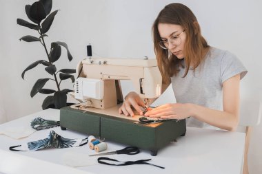 Gribi önlemek için evdeki dikiş makinesine koruyucu ilaç maskesi takan bir kız. Covid-19 beyaz arka planı olan odada.