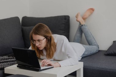 Evden Çalışan Çekici Genç Kadın - Kadın Girişimci, kanepede dizüstü bilgisayarla oturuyor ve Ev Konforu 'ndan cep telefonunu kontrol ediyor. uzak çalışıyor