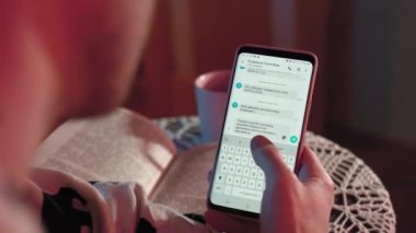 Bir adam telefonuyla yazı yazıyor ve renkli ışıklarla dolu bir odada oturuyor. Adam pijama giymiş. Arka planda bir kitap ve bir fincan çay var.