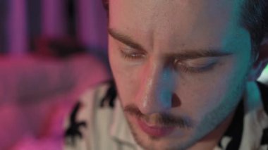 Bıyıklı ve sakallı bir adamın portresi aşağı bakıyor ve oturuyor. Pijama giyiyor ve odada pembe ve mavi renkler var. Arkasında bir kedi var.