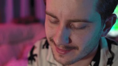 Bıyıklı ve sakallı bir adamın portresi aşağı bakıyor ve oturuyor. Pijama giyiyor ve odada pembe ve mavi renkli ışıklar var. Adam gülümsüyor ve gülüyor.