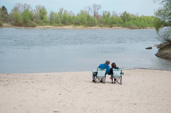 couple man and woman  sitting on riverside on chairs  in spring and watching film on device and resting on holidays, warm days