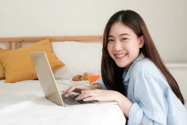 Genç Asyalı bir kadın laptopta çalışırken kameraya bakıyor ve kahvaltıyla birlikte parlak bir şekilde gülümsüyor. Ev konseptinden çalışıyor..