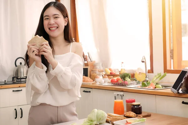 Tatlı Asyalı kadın sandviç yiyor ve mutfakta kahvaltı hazırlıyor..