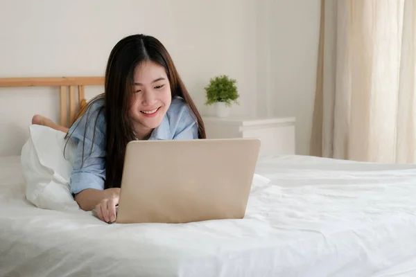 Mavi tişörtlü tatlı Asyalı kadın evde bilgisayarla çalışıyor. Yatak odasında çalışan gülümseyen kadın..