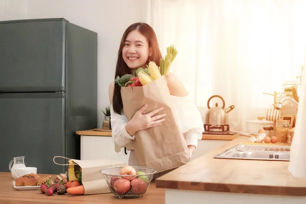 キッチンには果物や野菜の入ったショッピングバッグを持っている可愛いアジアの女性が立っていて カメラを見て明るい笑顔で — ストック写真