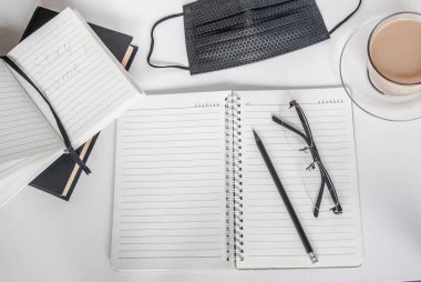 Siyah tıbbi maske ve defter. Bir fincan kahve ve bir kalemin yanında. Evden çalışıyorum. Karantinada evinizde kalın. Coronavirus konsepti /
