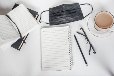 Siyah tıbbi maske ve defter. Bir fincan kahve ve bir kalemin yanında. Evden çalışıyorum. Karantinada evinizde kalın. Coronavirus konsepti /