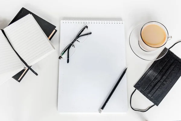 Siyah tıbbi maske ve defter. Bir fincan kahve ve bir kalemin yanında. Evden çalışıyorum. Karantinada evinizde kalın. Coronavirus konsepti /