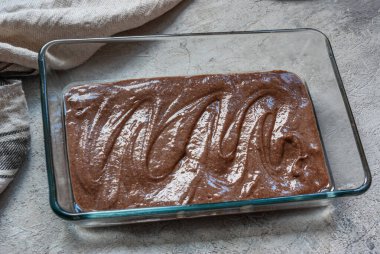 Kek hamuru, bej bir havlunun yanındaki gri beton masada, dikdörtgen bir cam kabın içinde. Yemek pişirme süreci