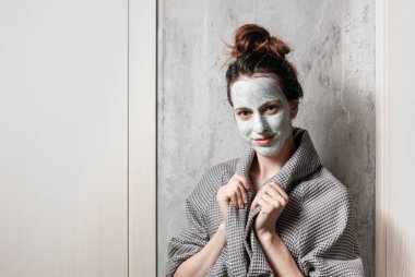 Kaplıcada, gri bornozlu bir kız evinde gri-bej duvarlı bir yüz maskesi yapıyor.