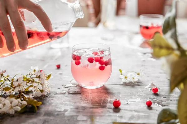 El, çiçekli ve yapraklı gri eski püskü bir masanın üzerindeki bir bardağa limonata döker.