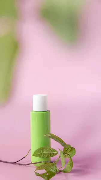 Green bottle with a skin care product next to fresh leaves on a pink background. Vertical copyspace beauty concept.