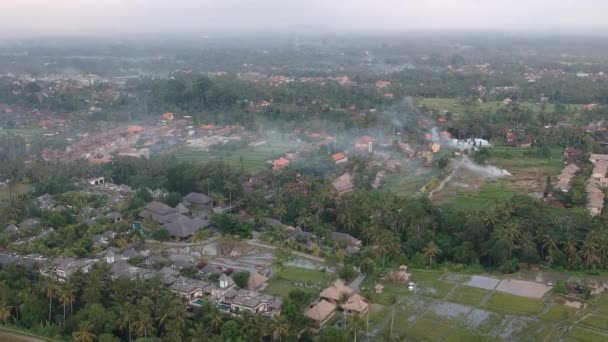 Muñeca de drones sobre la selva de Ubud en Bali, palmeras y casas con techos marrones y grises son visibles, una hoguera está fumando. clima nublado y gris — Vídeo de stock