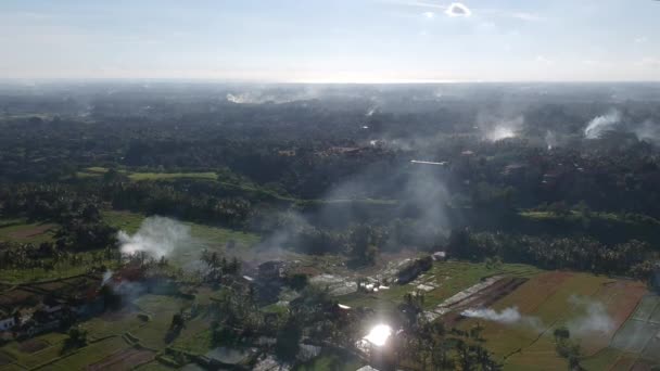 El quadrocopter aterriza verticalmente, disparando paralelamente al suelo, el humo de los incendios y el resplandor del sol son visibles en el reflejo del agua en los campos de arroz en Ubud Bali — Vídeo de stock