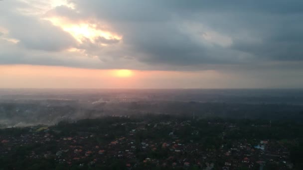 Il quadrocopter vola sopra Ubud a Bali, sono visibili incendi con fumo e tetti rossi delle case, si allontana dal tramonto, tutto il cielo è in nuvole e il sole giallo-arancio è visibile in — Video Stock