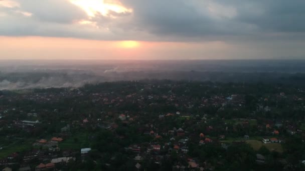 El quadrocopter despega sobre Ubud en Bali, el camino de los artistas y los tejados rojos de las casas son visibles, se aleja de la puesta del sol, todo el cielo está en las nubes y el sol amarillo-naranja es visible — Vídeos de Stock