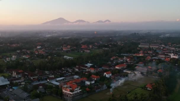 Hermosa puesta de sol con un dron volando sobre Ubud Bali, y muchas casas con techos rojos, montañas en el horizonte y un globo con nubes pintorescas y cielo azul — Vídeo de stock