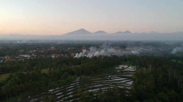 Um drone voando de volta sobre Ubud Bali, e muitas casas com telhados vermelhos, uma fogueira de fumar à noite, montanhas no horizonte escondido atrás de nuvens — Vídeo de Stock