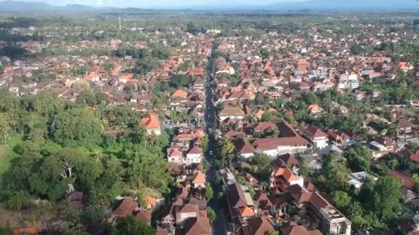 Drone volando sobre el centro de ubud jl.Raya Ubud en tiempo soleado, casas con techos brillantes, selva de palmas y montañas en el horizonte son visibles — Vídeos de Stock