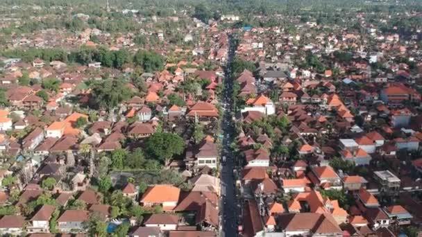 Quadrocopter volando sobre el centro de ubud jl.Raya Ubud en tiempo soleado, casas con techos rojos, selva de palmas y montañas en el horizonte son visibles — Vídeos de Stock