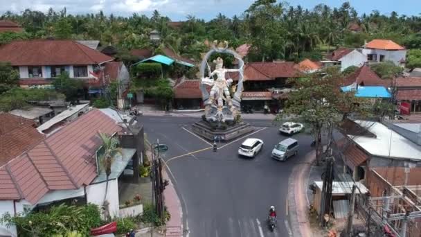 Volando por encima de la estatua de ARJUNA, pasando coches y scooters son visibles en la intersección de COK Gede Rai y Raya Ubud bali en tiempo soleado — Vídeos de Stock